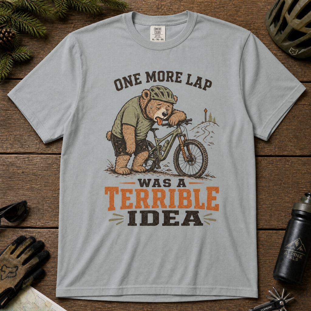 Granite gray t-shirt with bear on bike graphic and text on wooden surface with outdoor gear.