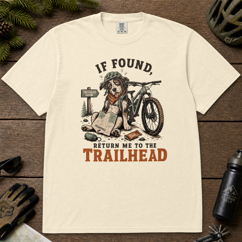 Ivory white t-shirt with a graphic of a dog on a bike and text 'If Found, Return Me to the Trailhead' on a wooden surface with outdoor gear.