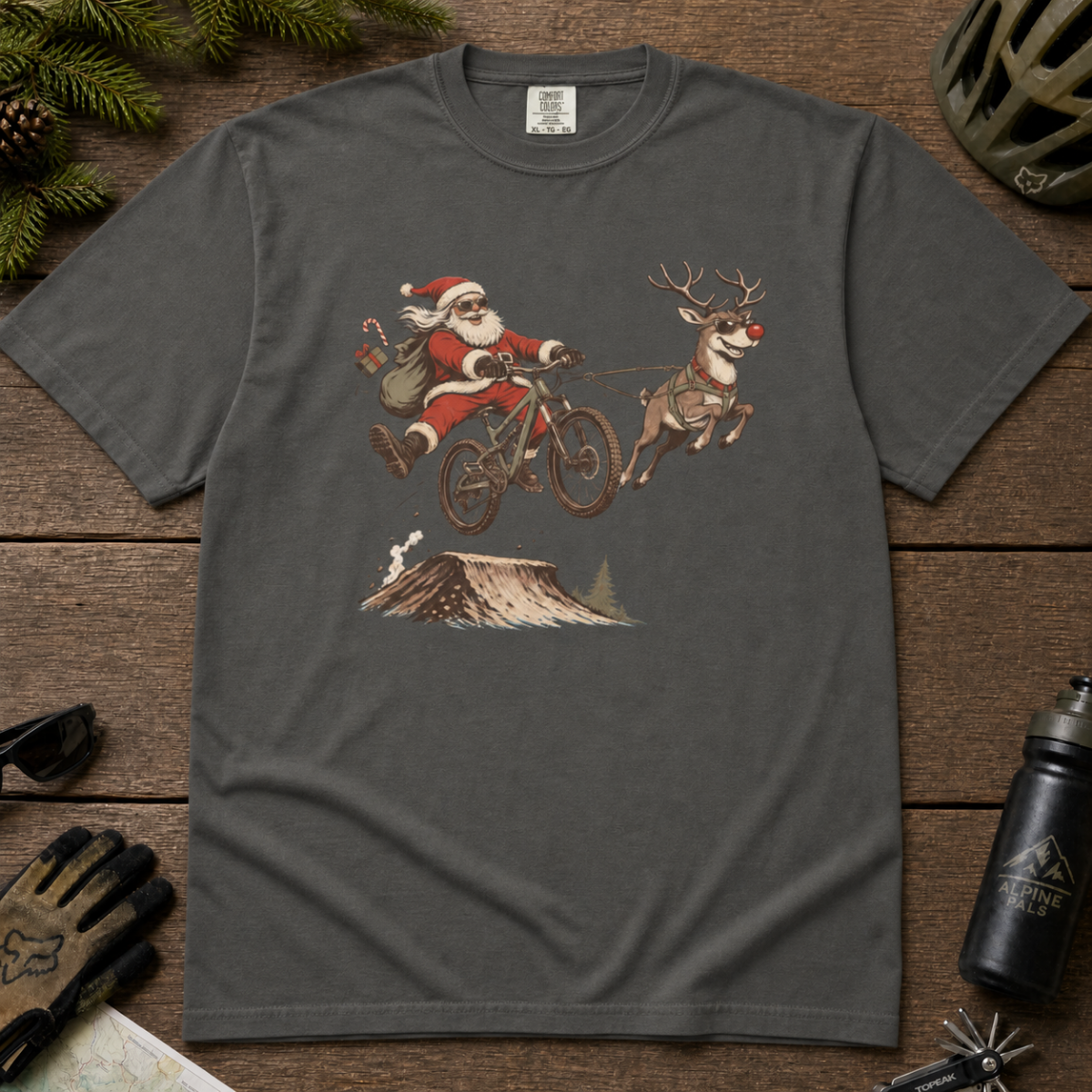 Pepper gray t-shirt with a graphic of Santa Claus riding a bike with a reindeer, on a wooden surface with outdoor gear.