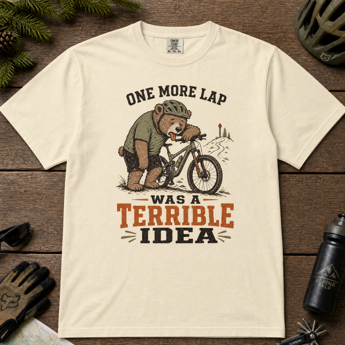 Ivory white t-shirt with bear graphic and text on a wooden surface with cycling accessories.