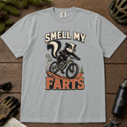 Granite gray t-shirt with a graphic of a skunk on a bike and text 'Smell My Farts' on a wooden surface.