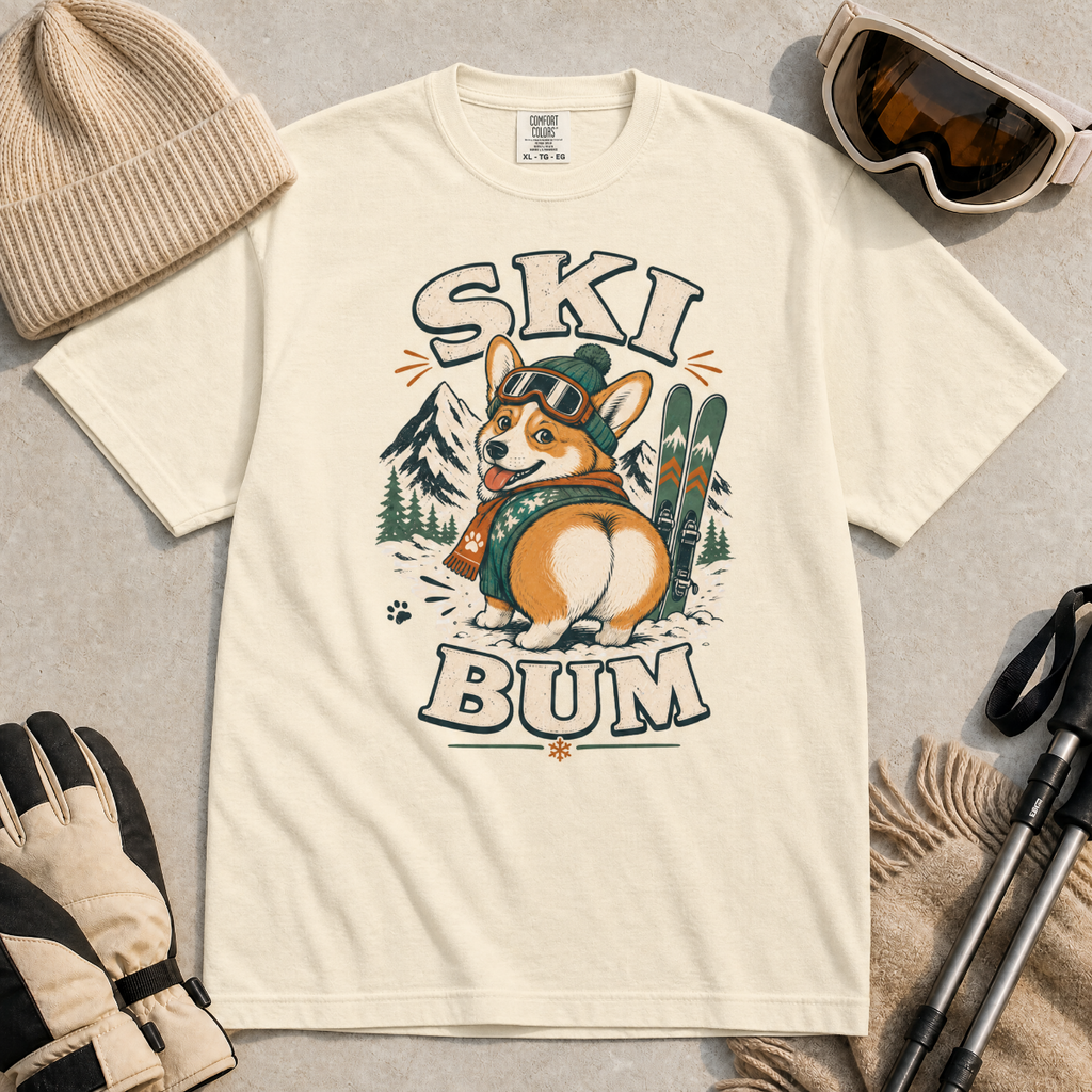 Ivory white t-shirt with a corgi graphic and 'Ski Bum' text, surrounded by winter accessories on a gray surface.