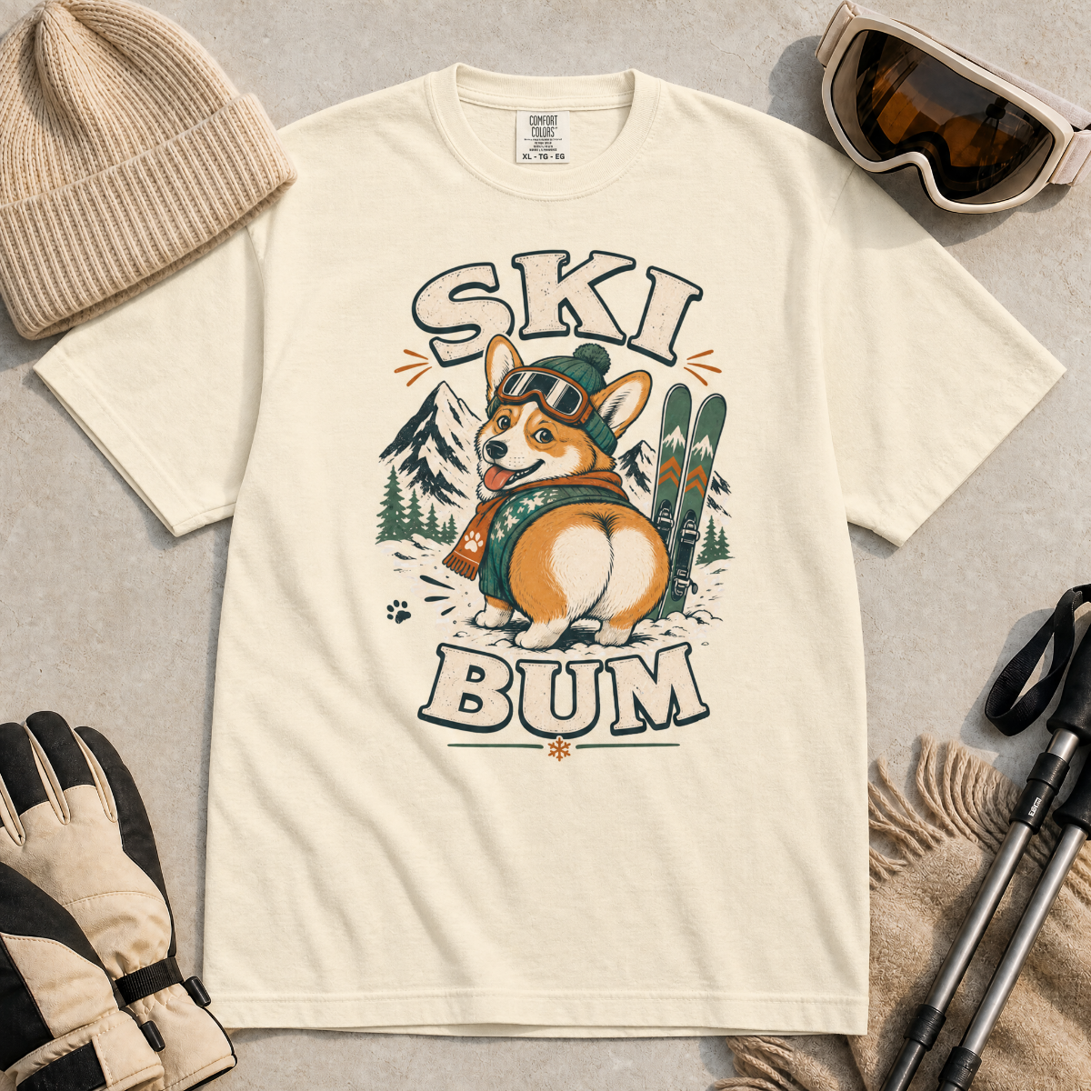Ivory white t-shirt with a corgi graphic and 'Ski Bum' text, surrounded by winter accessories on a gray surface.