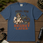 Midnight blue t-shirt with a dog and bicycle graphic and 'Pedal Now Regret Later' text on a wooden surface with cycling accessories.
