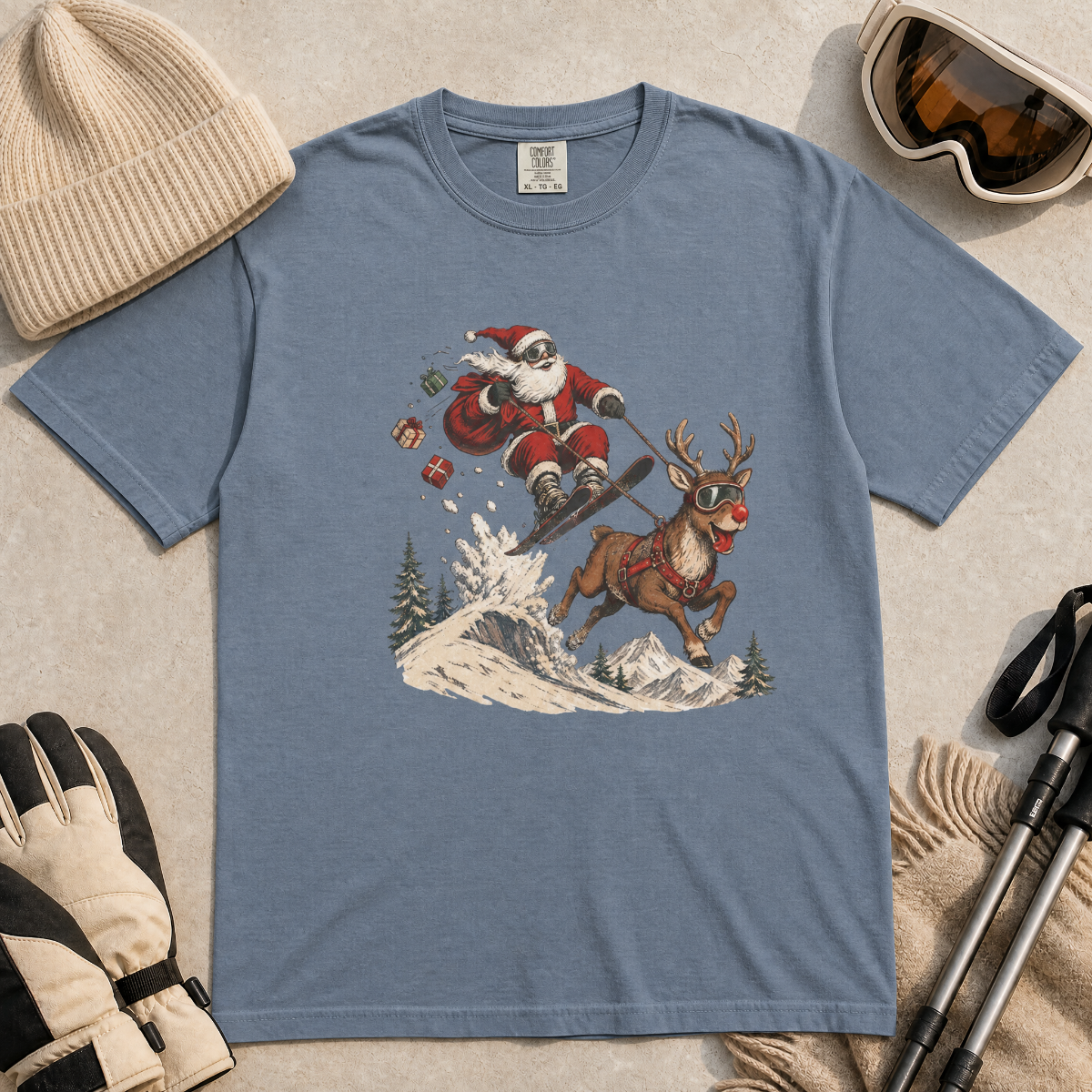Blue jean colored t-shirt with Santa Claus and reindeer graphic, surrounded by winter accessories on a beige surface.