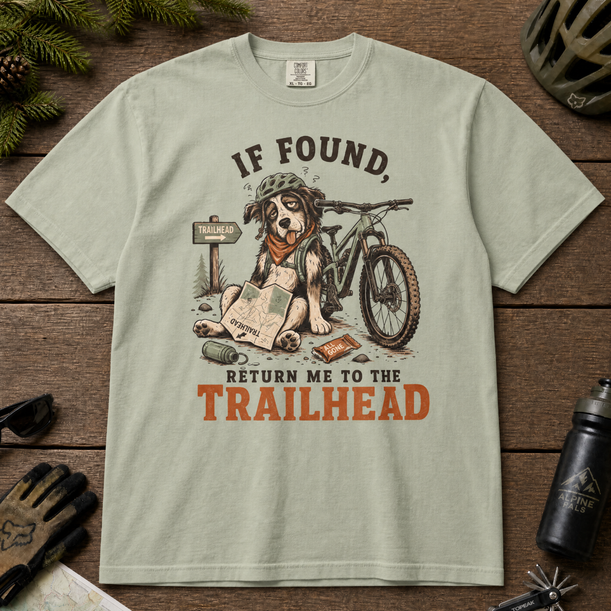 Bay green t-shirt with a dog and bicycle graphic and text 'If found, return me to the trailhead' on a wooden surface.