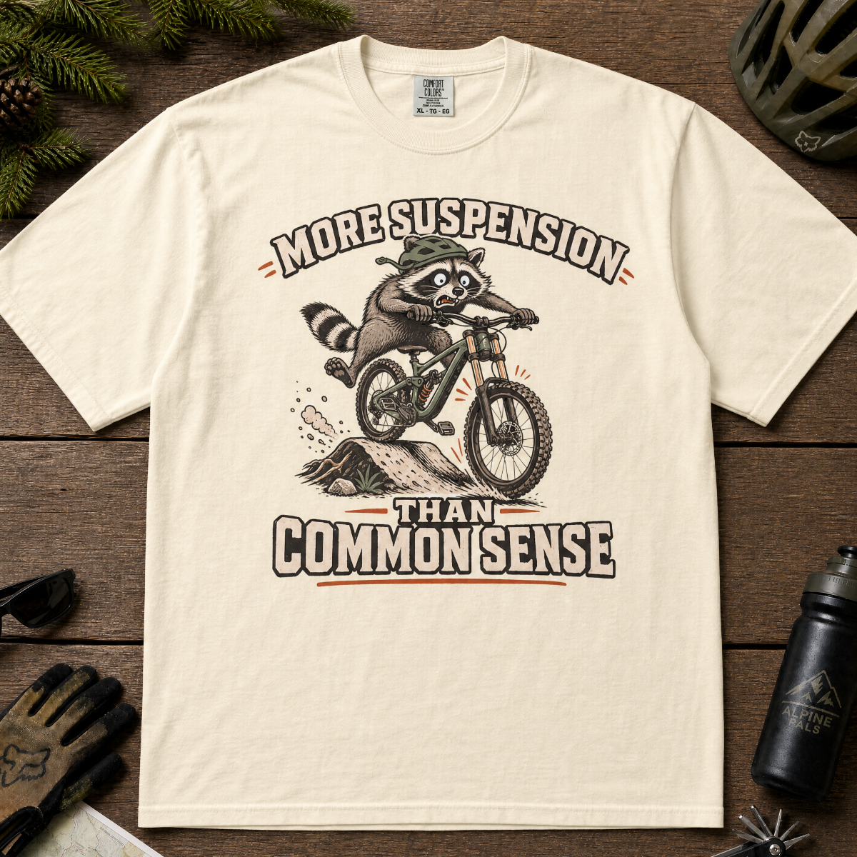 Ivory white t-shirt with a raccoon riding a bike graphic and text 'More suspension than common sense'.