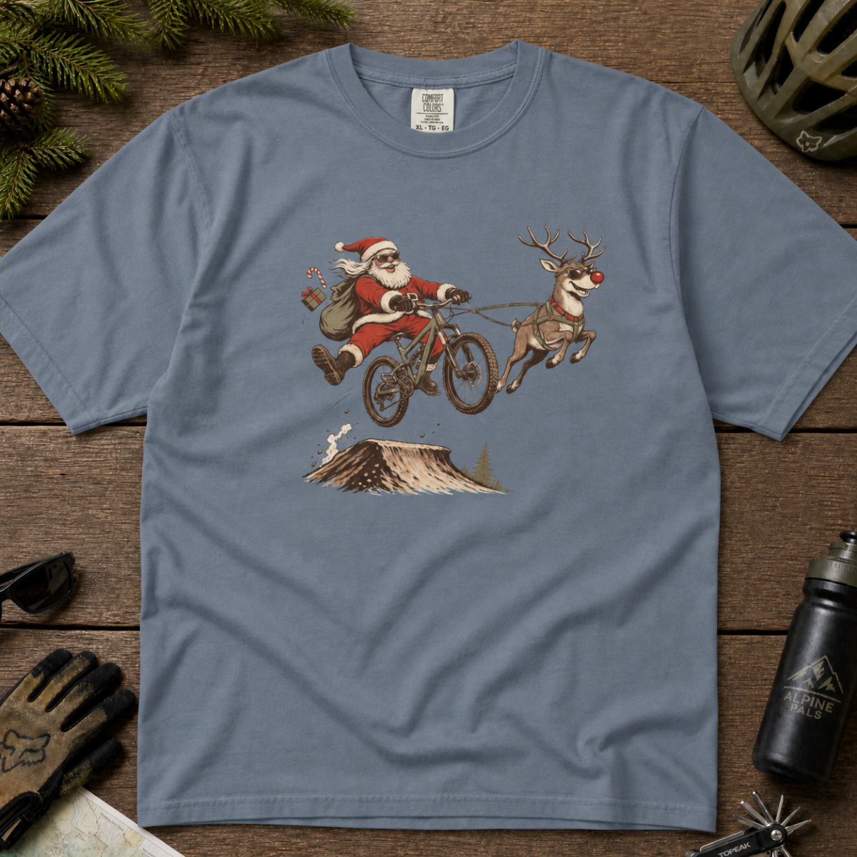 Blue jean colored t-shirt with Santa Claus riding a bike and reindeer design on a wooden surface.