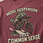 Brick red t-shirt with a raccoon riding a bike graphic and text 'More suspension than common sense'.