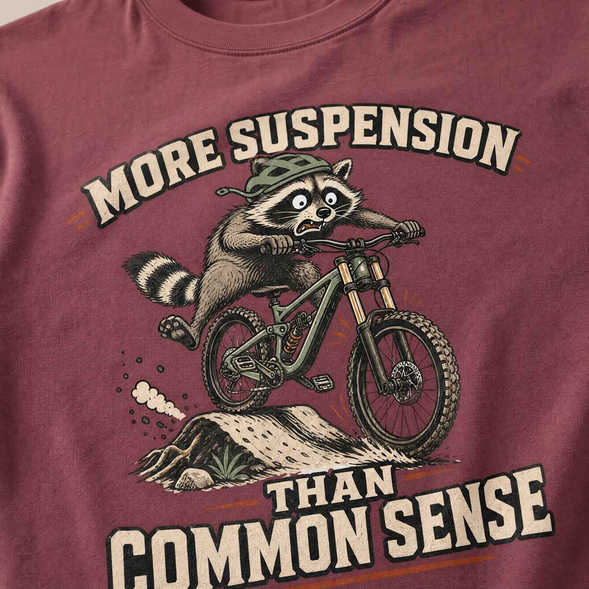 Brick red t-shirt with a raccoon riding a bike graphic and text 'More suspension than common sense'.