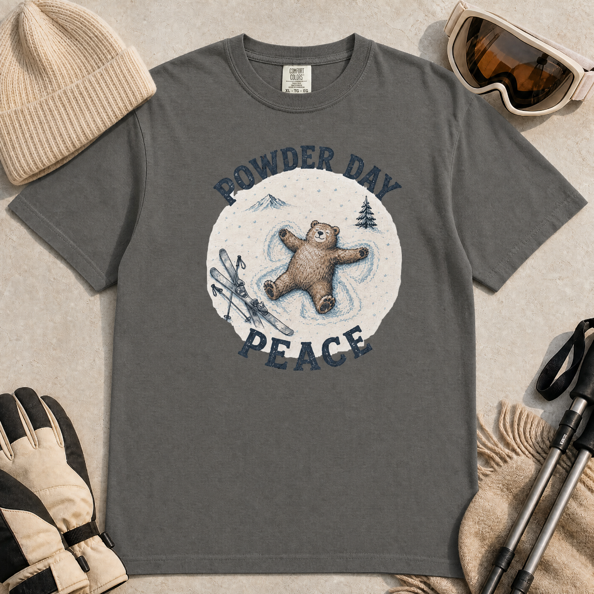 Flat lay of a pepper gray t-shirt featuring a peaceful bear snow angel ski graphic with “Powder Day Peace” text, styled with winter accessories.