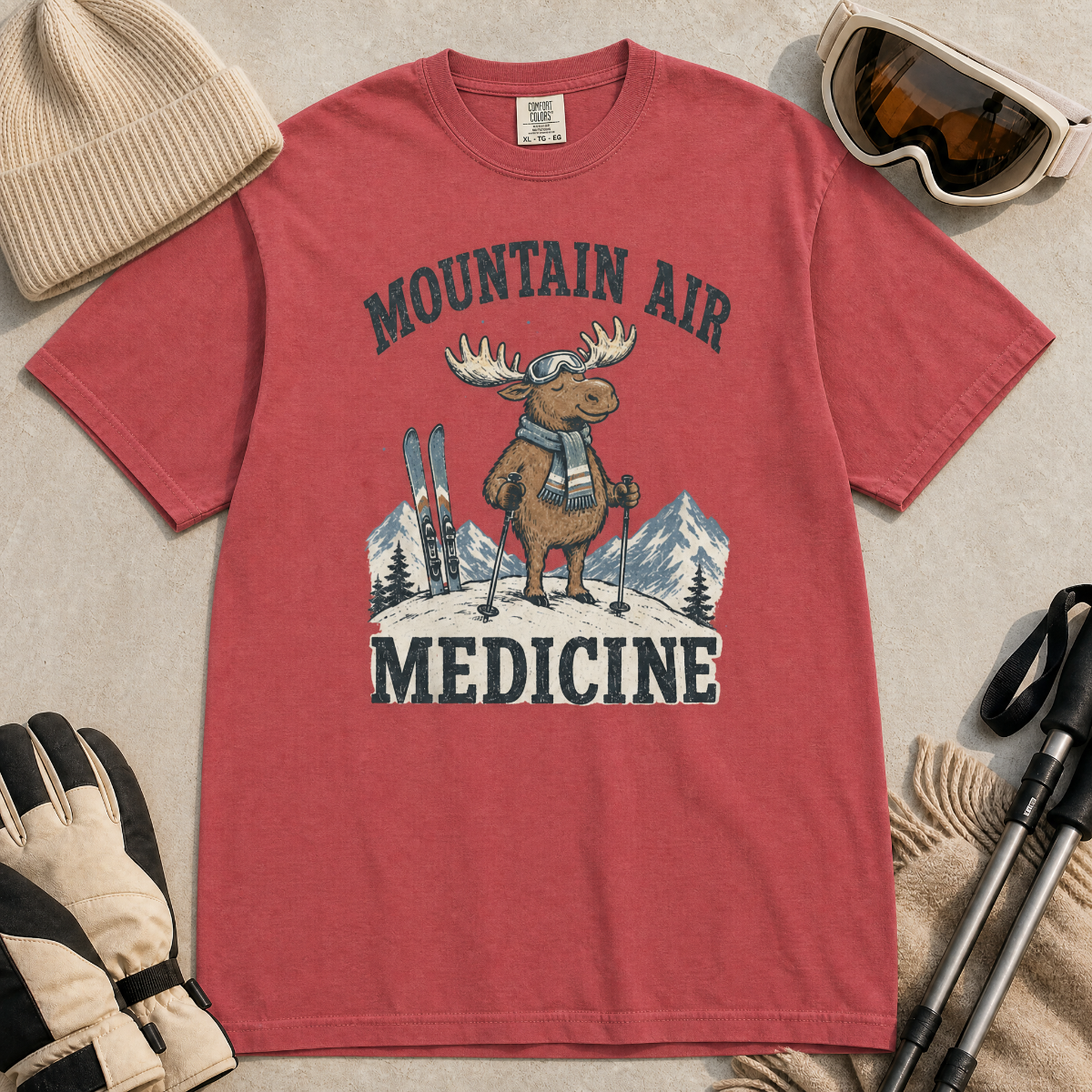 Flat lay of a crimson red t-shirt featuring a peaceful moose ski graphic with “Mountain Air Medicine” text, styled with winter accessories.