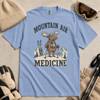 Flat lay of a washed denim blue t-shirt featuring a peaceful moose ski graphic with “Mountain Air Medicine” text, styled with winter accessories.