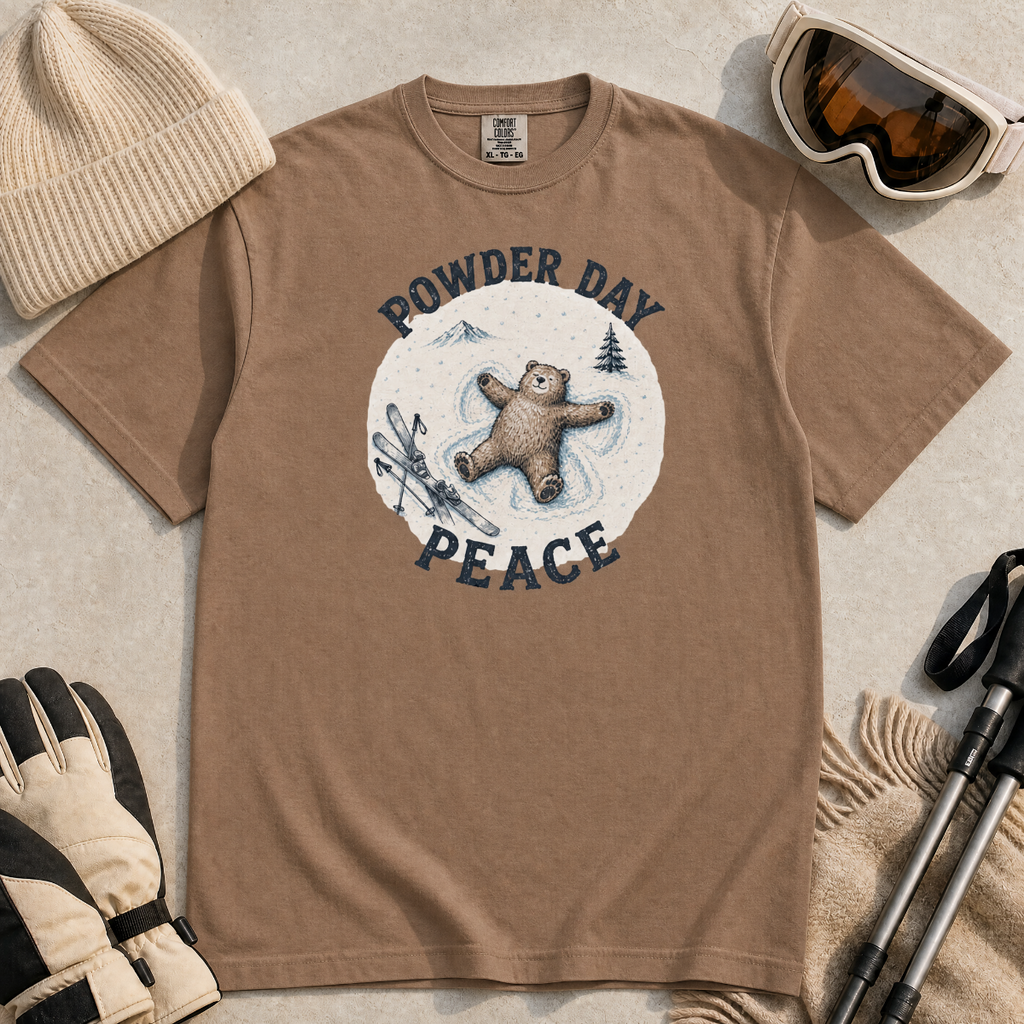 Flat lay of an espresso brown t-shirt featuring a peaceful bear snow angel ski graphic with “Powder Day Peace” text, styled with winter accessories.