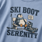 Close-up of a washed denim blue t-shirt with a vintage ski boot-room design showing a relaxed penguin on a bench beside ski boots, with “Ski Boot Serenity” text.