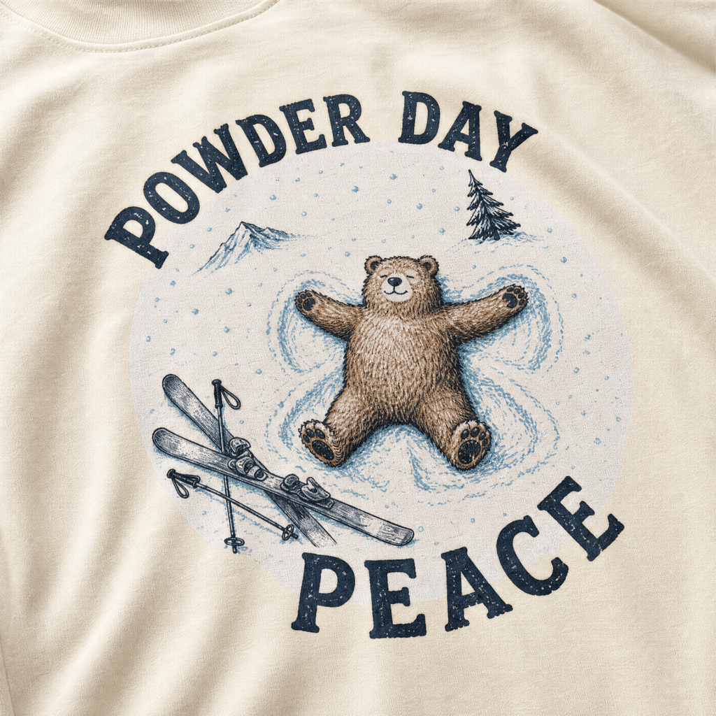 Close-up of an ivory white t-shirt with a vintage ski design showing a relaxed bear making a snow angel beside skis, with “Powder Day Peace” text.