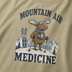 Close-up of a khaki colored t-shirt with a retro ski design showing a calm moose with goggles, scarf, ski poles, and mountain scenery, with “Mountain Air Medicine” text.