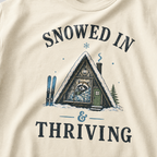 Close-up of an ivory white t-shirt with a vintage ski cabin design showing a cozy raccoon wrapped in a blanket inside an A-frame cabin, with “Snowed In & Thriving” text.