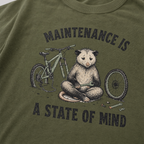 Hemp green t-shirt featuring a possum meditating while fixing a bike with text, "Maintenance is a State of Mind".