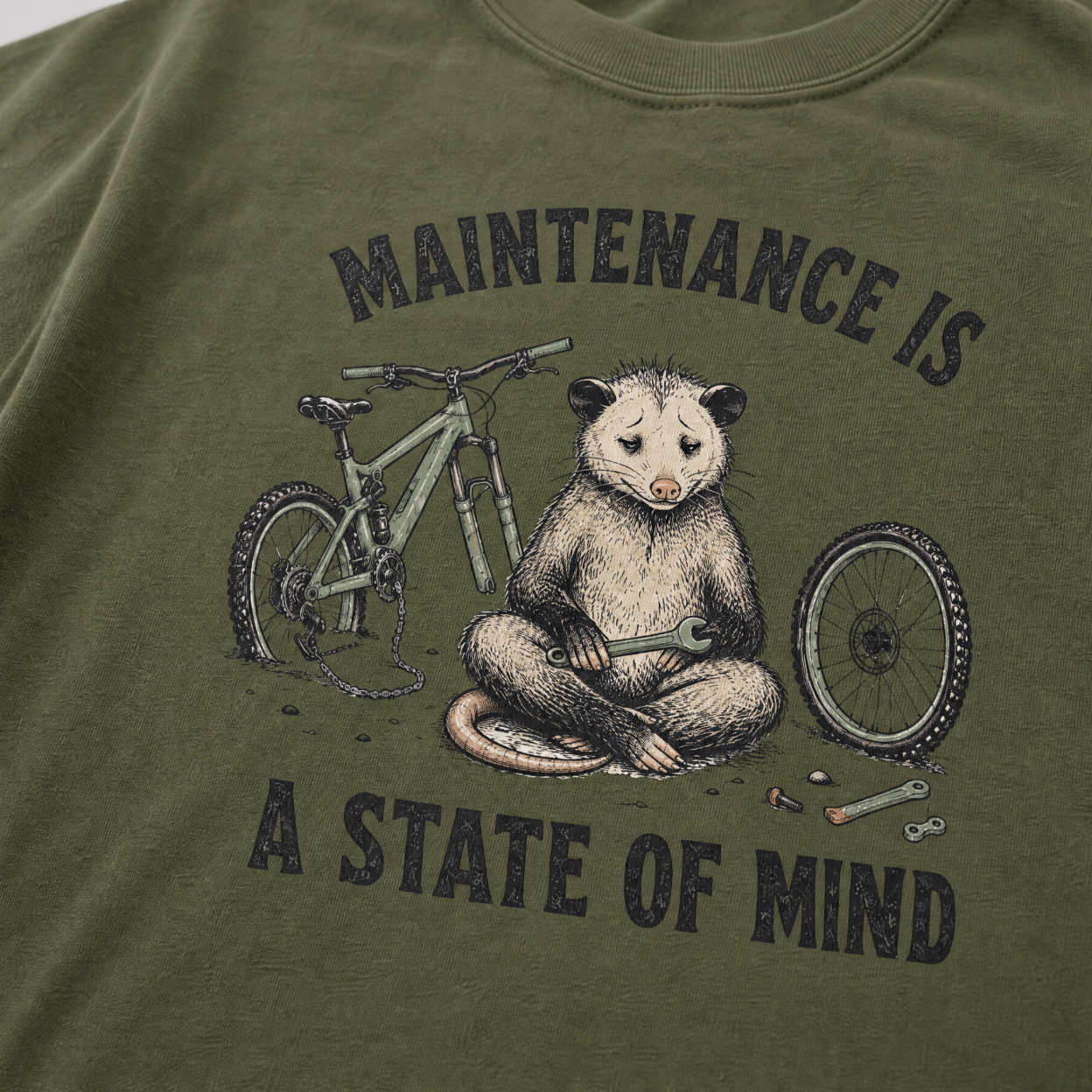 Hemp green t-shirt featuring a possum meditating while fixing a bike with text, "Maintenance is a State of Mind".
