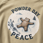 Close-up of a bay green t-shirt with a vintage ski design showing a relaxed bear making a snow angel beside skis, with “Powder Day Peace” text.