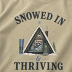 Close-up of a bay green t-shirt with a vintage ski cabin design showing a cozy raccoon wrapped in a blanket inside an A-frame cabin, with “Snowed In & Thriving” text.
