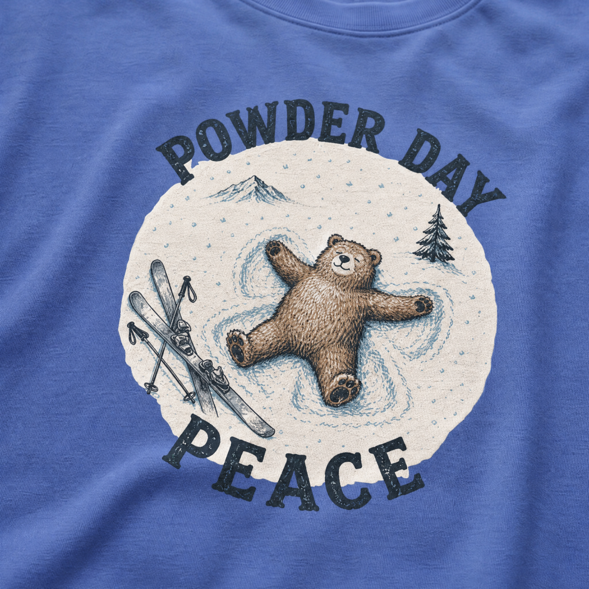 Close-up of a mystic blue t-shirt with a vintage ski design showing a relaxed bear making a snow angel beside skis, with “Powder Day Peace” text.