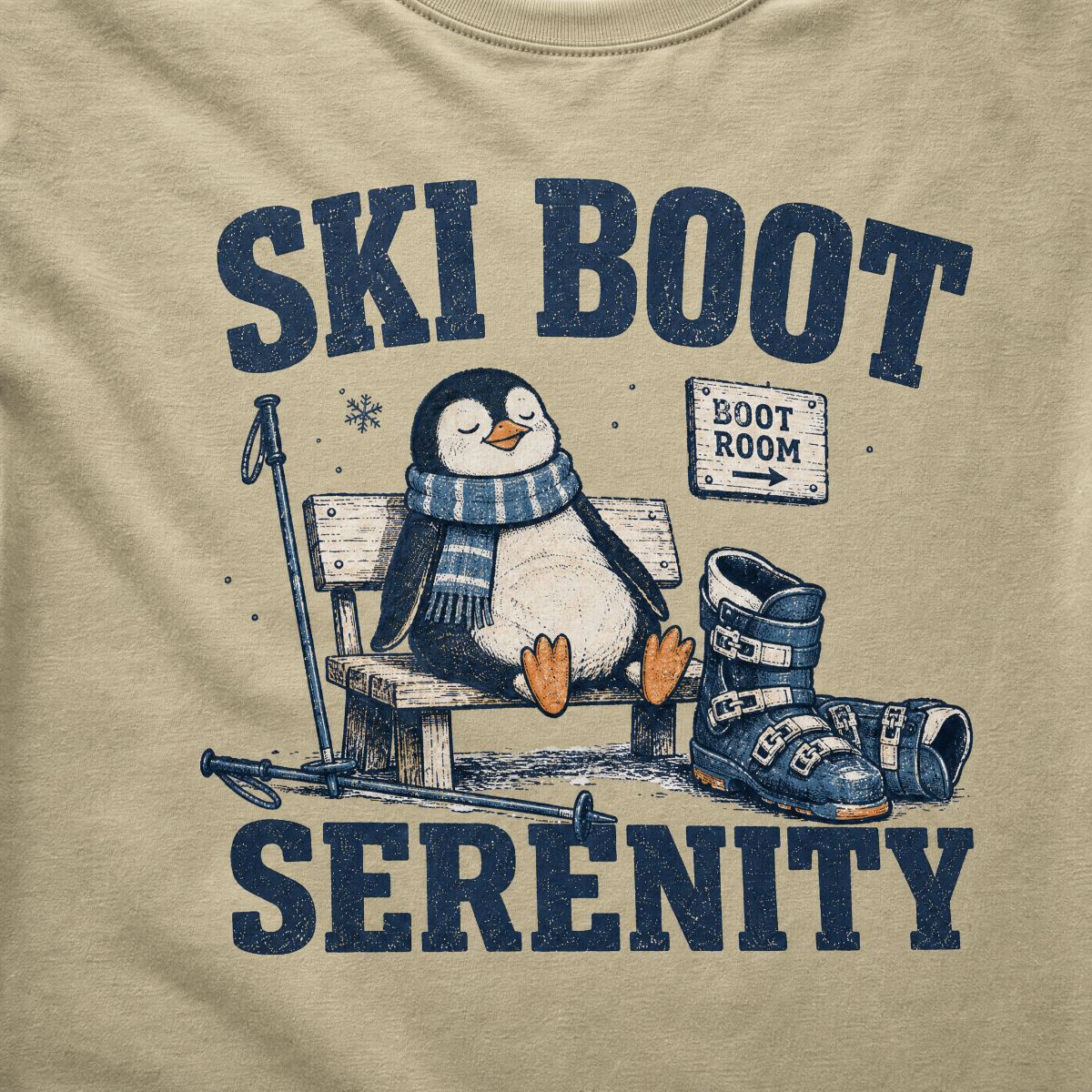 Close-up of a khaki colored t-shirt with a vintage ski boot-room design showing a relaxed penguin on a bench beside ski boots, with “Ski Boot Serenity” text.