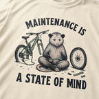 Ivory white t-shirt featuring a possum meditating while fixing a bike with text, "Maintenance is a State of Mind".