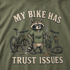 Moss green t-shirt with a raccoon graphic and bicycle illustration, featuring text 'My bike has trust issues'.