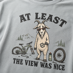 Granite gray t-shirt of a goat holding a bicycle pedal with a broken mountain bike surrounding it. Included text, "At Least the View Was Nice".