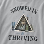 Close-up of a granite gray t-shirt with a vintage ski cabin design showing a cozy raccoon wrapped in a blanket inside an A-frame cabin, with “Snowed In & Thriving” text.