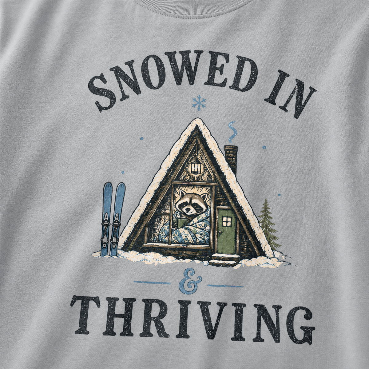 Close-up of a granite gray t-shirt with a vintage ski cabin design showing a cozy raccoon wrapped in a blanket inside an A-frame cabin, with “Snowed In & Thriving” text.