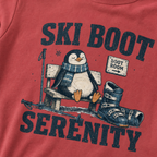 Close-up of a crimson red t-shirt with a vintage ski boot-room design showing a relaxed penguin on a bench beside ski boots, with “Ski Boot Serenity” text.