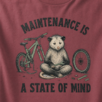 Brick red t-shirt featuring a possum meditating while fixing a bike with text, "Maintenance is a State of Mind".