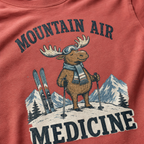 Close-up of a crimson red t-shirt with a retro ski design showing a calm moose with goggles, scarf, ski poles, and mountain scenery, with “Mountain Air Medicine” text.