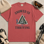 Flat lay of a crimson red t-shirt featuring a retro “Snowed In & Thriving” raccoon cabin graphic, styled with winter accessories like ski goggles, gloves, and poles.