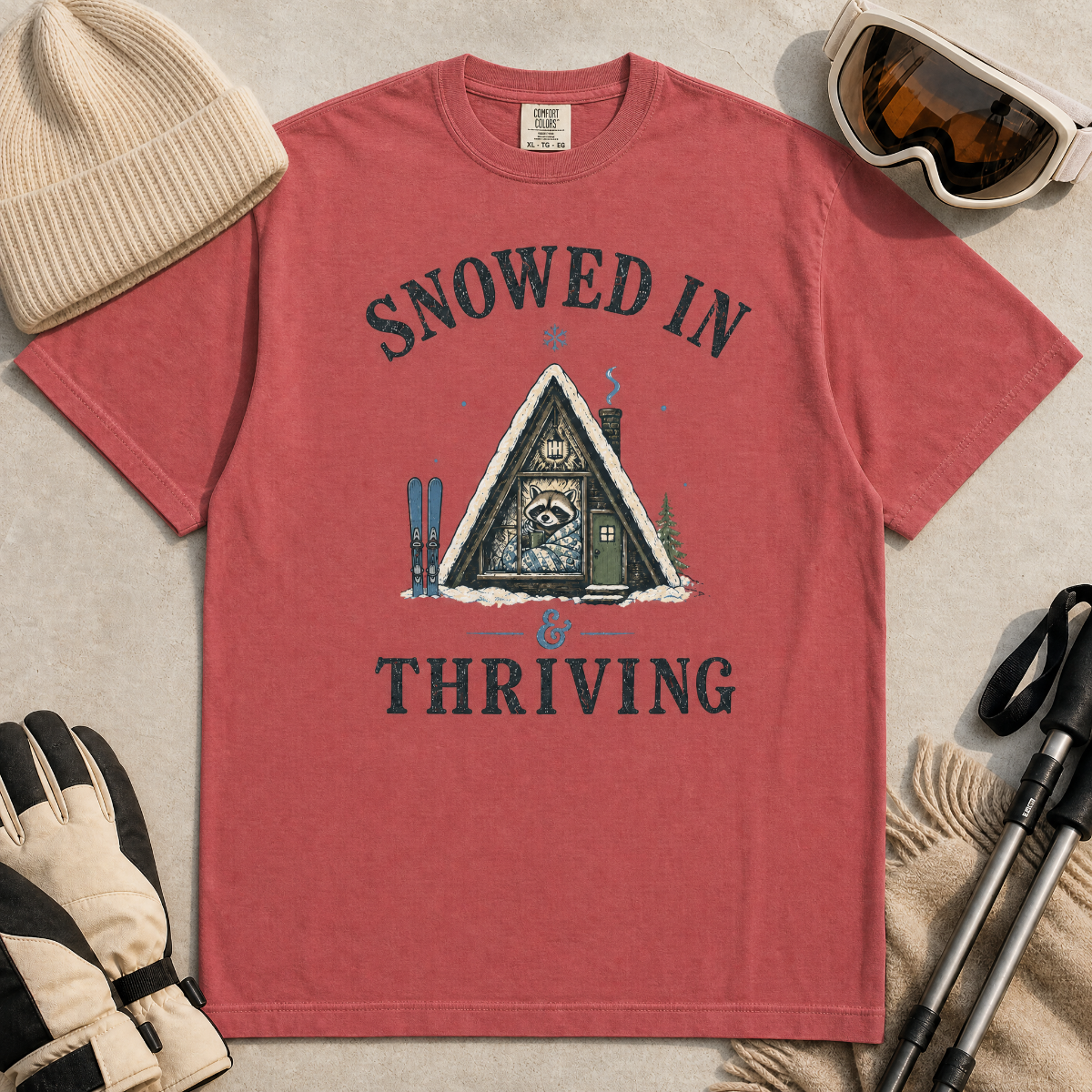 Flat lay of a crimson red t-shirt featuring a retro “Snowed In & Thriving” raccoon cabin graphic, styled with winter accessories like ski goggles, gloves, and poles.