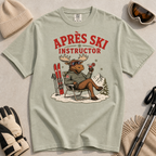 T-shirt with 'Après Ski Instructor' design featuring a moose, surrounded by skiing accessories on a light background.