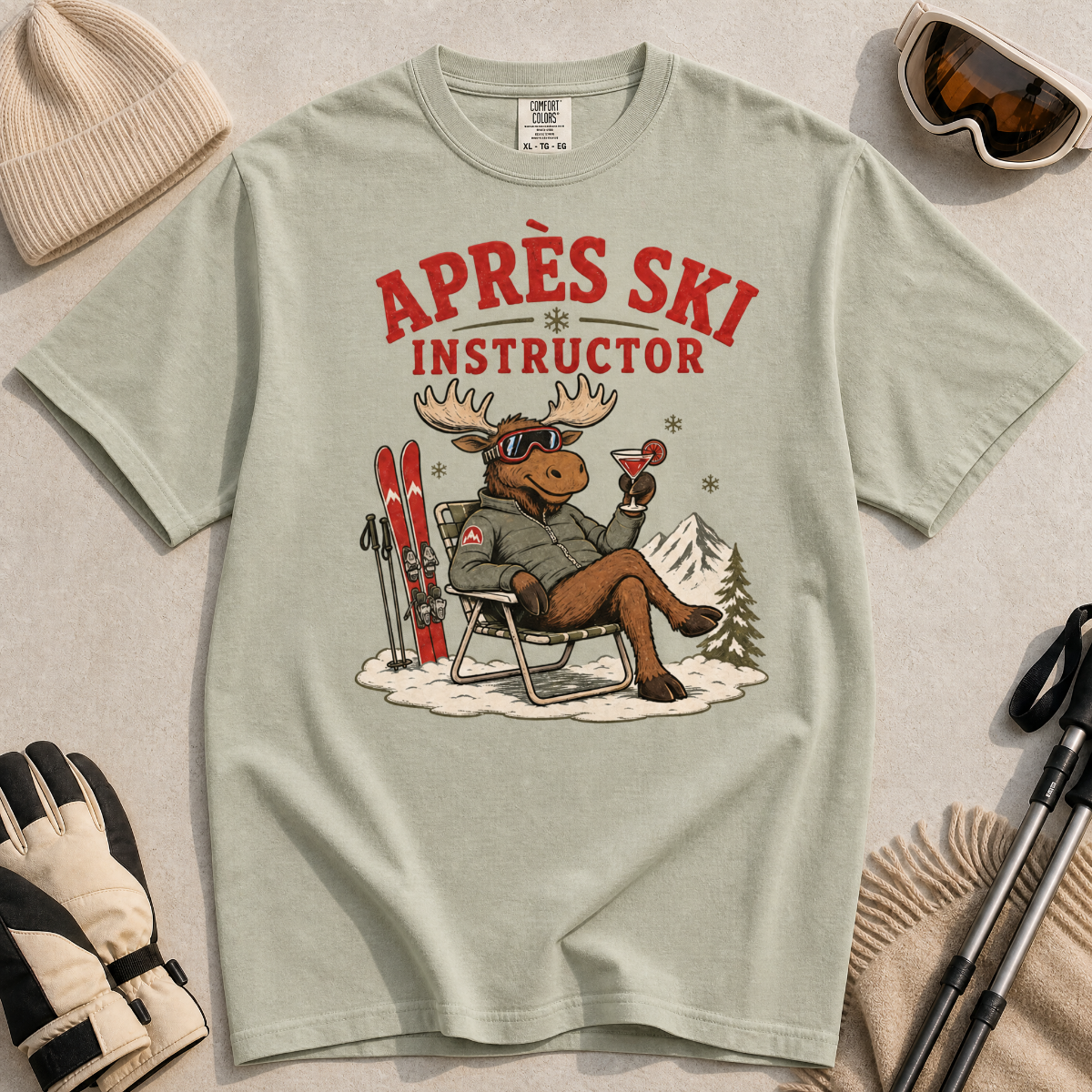 T-shirt with 'Après Ski Instructor' design featuring a moose, surrounded by skiing accessories on a light background.
