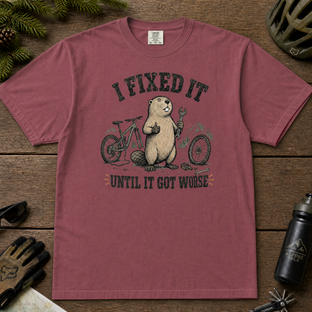 Brick red t-shirt with graphic of a beaver fixing a bike, text 'I FIXED IT UNTIL IT GOT WORSE', on a wooden surface with cycling accessories.