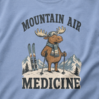 Close-up of a washed denim blue t-shirt with a retro ski design showing a calm moose with goggles, scarf, ski poles, and mountain scenery, with “Mountain Air Medicine” text.
