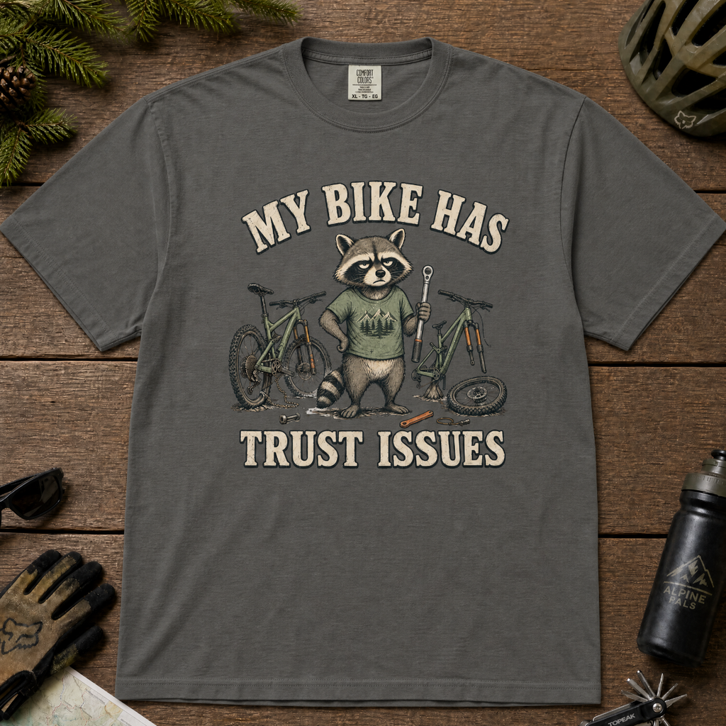 Pepper gray t-shirt with a raccoon graphic and text 'My bike has trust issues' on a wooden surface.