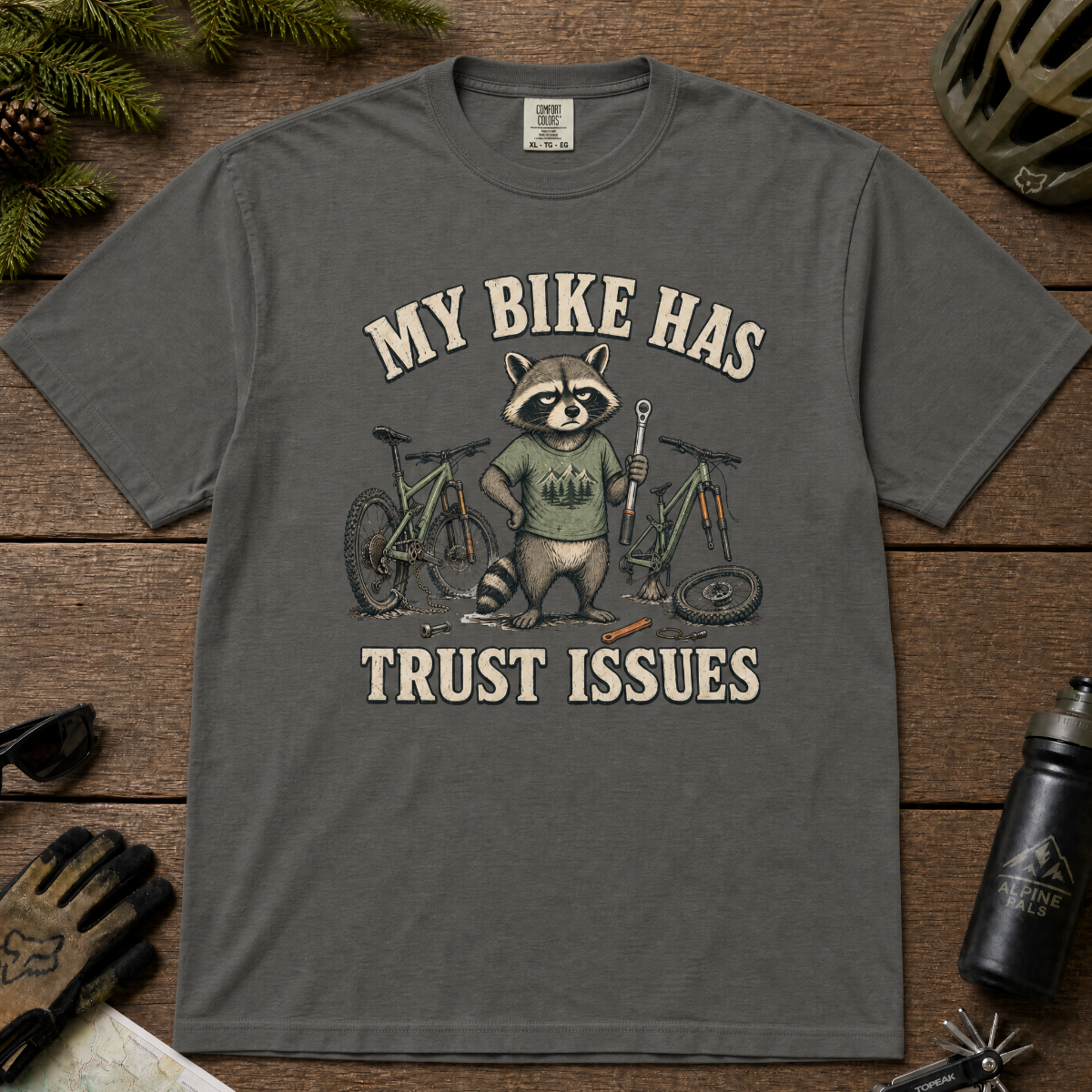 Pepper gray t-shirt with a raccoon graphic and text 'My bike has trust issues' on a wooden surface.