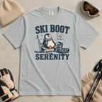 Flat lay of a granite gray t-shirt featuring a retro “Ski Boot Serenity” penguin design, styled with winter accessories like ski goggles, gloves, and poles.