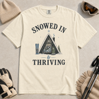 Flat lay of an ivory white t-shirt featuring a retro “Snowed In & Thriving” raccoon cabin graphic, styled with winter accessories like ski goggles, gloves, and poles.
