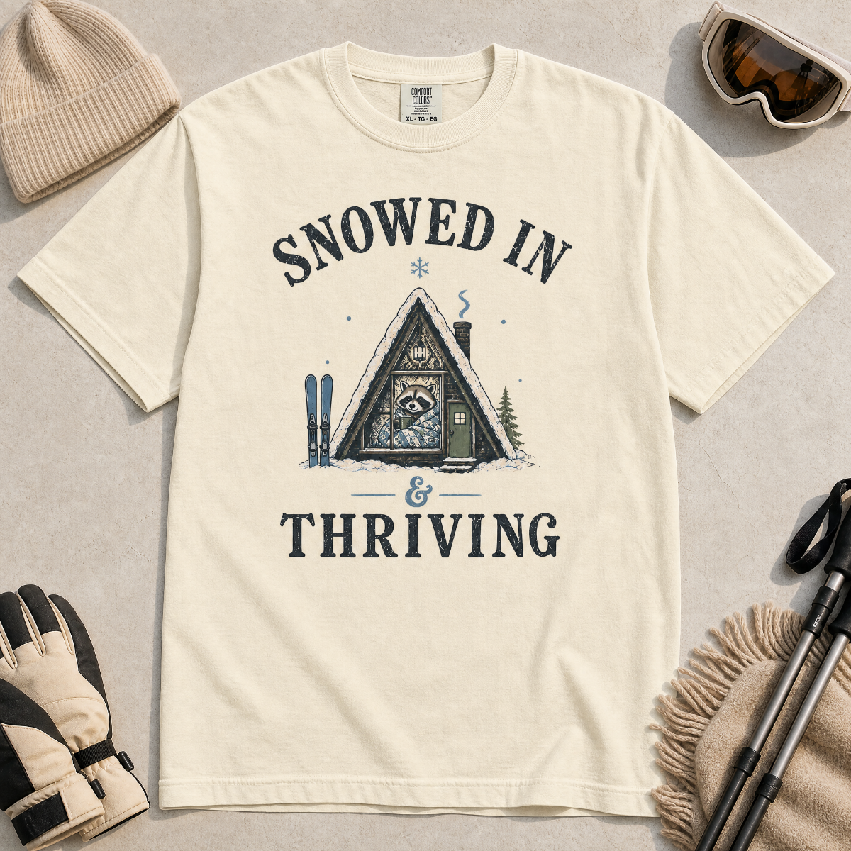Flat lay of an ivory white t-shirt featuring a retro “Snowed In & Thriving” raccoon cabin graphic, styled with winter accessories like ski goggles, gloves, and poles.