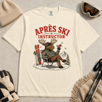 Ivory white t-shirt with a moose graphic and 'Après Ski Instructor' text, surrounded by skiing accessories on a light gray background.