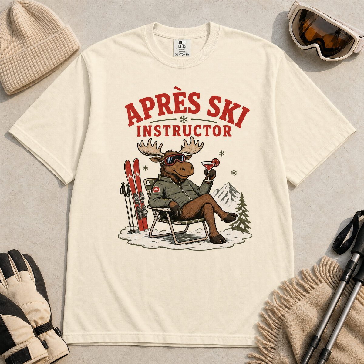 Ivory white t-shirt with a moose graphic and 'Après Ski Instructor' text, surrounded by skiing accessories on a light gray background.