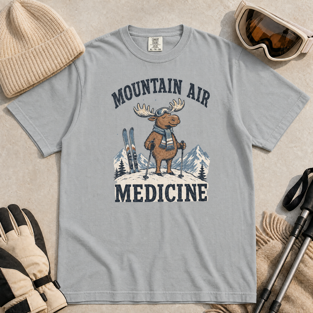 Flat lay of a granite gray t-shirt featuring a peaceful moose ski graphic with “Mountain Air Medicine” text, styled with winter accessories.